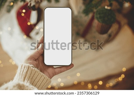 Hand holding smartphone with empty screen against stylish festive christmas gifts and golden lights. Christmas phone mock up. Space for text. Christmas advertising, smartphone app template