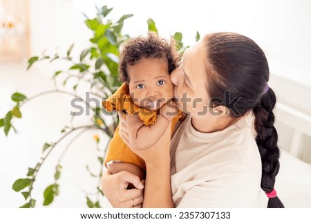 white Caucasian mom hugs and kisses an African-American black foster baby girl at home on the bed, maternal love and care, a mother of European appearance with a black child in her arms.