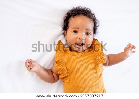 a laughing little African-American baby girl in an orange bodysuit on the bed at home, a funny six-month-old black newborn baby lies on the back and rejoices. View from above. Royalty-Free Stock Photo #2357307217