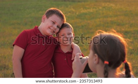 Cheerful little boys pose to girl taking photo with analog camera in twilight park. Happy little boys hug looking in sister camera lens in field. Brothers pose for photo to little girl in meadow