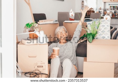 Happy senior woman involved in moving house sitting among cardboard boxes takes a break with a coffee, concept of moving, retirement, new life, buying, renting, apartment, house Royalty-Free Stock Photo #2357313537