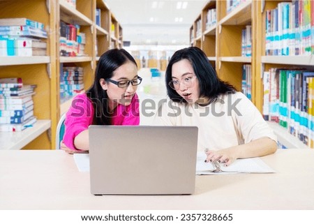 Smile young people looking laptop screen, communicating, two female students at the desk using working on laptop with book shelf in library background