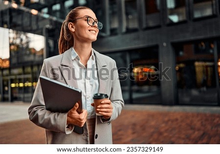Beautiful confident successful modern business woman wearing glasses holding a laptop and coffee and looking to the side while standing outside in the evening behind a dark blurred background.