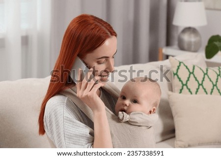 Mother talking on smartphone while holding her child in sling (baby carrier) at home
