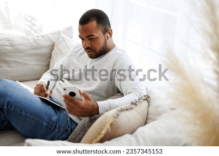 African american man in earphone holding smartphone during online courses and writing on notebook on couch at home Royalty-Free Stock Photo #2357345153