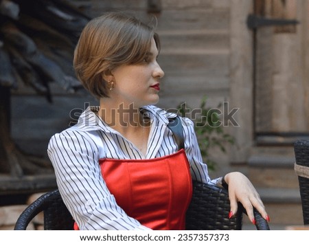 A stylish beautiful girl in fashionable clothes with a white shirt and a red top is sitting in the city near a wooden wall.