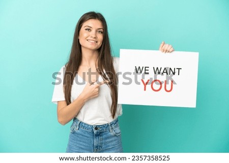 Young caucasian woman isolated on blue background holding We Want You board and  pointing it