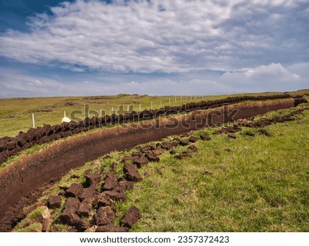 Freshly cut blocks of peat stacked in rows above a trench in the peat base and lying in the sun to dry. Taken in the Outer Hebrides, Scotland, UK on a sunny day in summer.  Royalty-Free Stock Photo #2357372423