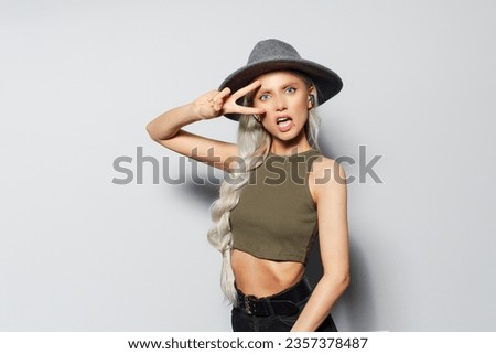 Studio portrait of attractive happy blonde girl with wireless earbuds in ears, showing peace sign, on white background. Wearing grey hat and green shirt.