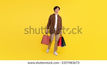 Sale, consumerism. Smiling guy after shopping many colored packages bags posing on yellow background, studio shot. Customer standing with purchases advertising offer. Panorama, full length