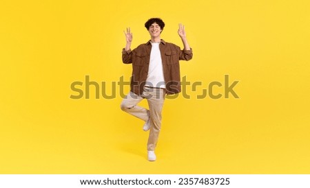 Keep calm. Young man meditating standing on one leg, making om or okay gesture posing over yellow background in studio. Yoga meditation exercise and mindfulness concept. Panorama, full length shot
