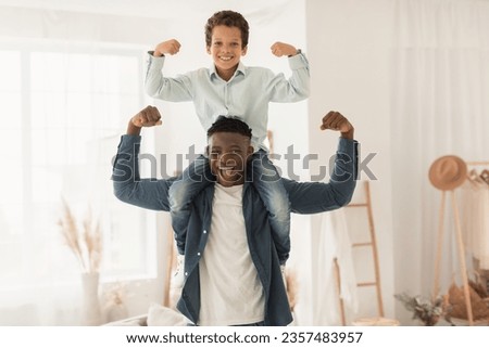 Raising Little Champion. Black Stepdad Holding His Son On Neck Showing Their Biceps Muscles At Home, Daddy And Kid Boy Demonstrating Their Strength And Power. We Are Strong, Leadership in Child Royalty-Free Stock Photo #2357483957