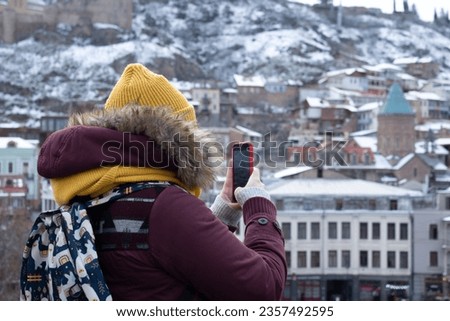 View from back. Woman traveler takes pictures on red mobile phone of landscape with view of winter Georgian city of Tbilisi. Concept of tourism, adventure, travel. Smartphone. Mountain, house, church