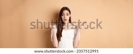 Surprised caucasian girl look with disbelief and amazement, spread hands sideways and gasping confused, cant understand what happening, know nothing, standing on beige background. Royalty-Free Stock Photo #2357512991
