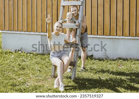Married couple with an infant child sits on wooden ladder near rural country house. Young family with an infant near wooden house, portrait photography.