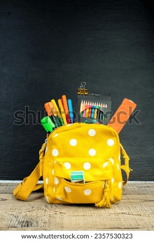 Yellow backpack with white polka dots with different colorful stationery on table. Black background. Back to school