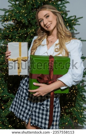 Young carefree woman with blond hair dressed in a skirt and blouse holding gifts on the background of the Christmas tree.