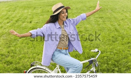 Happy beautiful curly Spanish  woman in a straw hat rides a bicycle without arms spread her arms to the side laughs with pleasure. Girl overcame fear rushes towards new adventures. Biking outdoors.