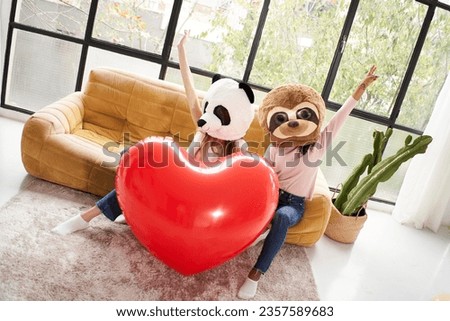 Couple of unrecognizable girls with stuffed animal masks holding giant red inflatable heart sitting on sofa. People in costume celebrating valentine day indoors. Panda bear and Sloth. Copy space.