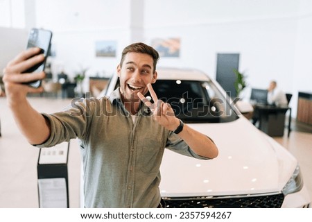 Portrait of happy buyer male taking selfie with victory sign on smartphone from auto dealership after bought new car. Smiling young man choosing new vehicle in showroom and making photo using phone.