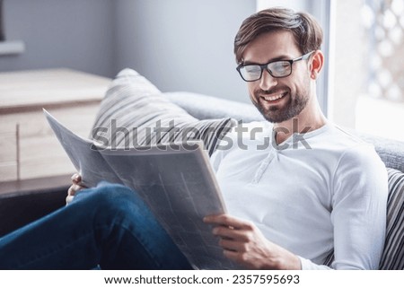 Handsome man in eyeglasses is reading a newspaper and smiling while sitting on couch at home