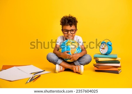 Full body photo of clever positive small kid sit floor hold world globe pile stack book clock notebook isolated on yellow color background