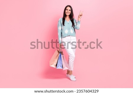 Full length photo of shiny sweet woman dressed blue cardigan showing v-sign holding shoppers isolated pink color background