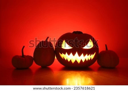 Halloween spooky angry jack-o-lantern and three little pumpkins silhouettes. Carved glowing pumpkin stands on surface in dark night. Back wall with copy space is highlighted red with shadows.