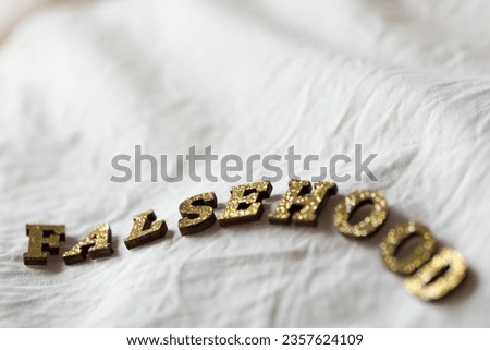 the word "falsehood" in metallic coated wooden letters on soft white cloth