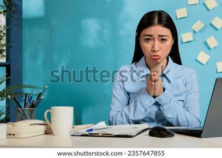 Professional woman asking for forgiveness by making pleading gesture with hands while working from work station in contemporary office. Employee sitting at desk with laptop asking for second chance.