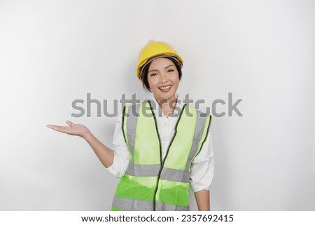 A smiling Asian woman labor wearing safety helmet and vest, pointing to copy space beside her, isolated by white background. Labor's day concept.