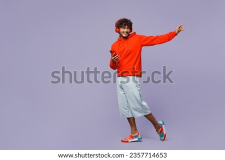 Full body young Indian man wear red orange hoody casual clothes listen to music in headphones use mobile phone isolated on plain pastel light purple color background studio portrait. Lifestyle concept