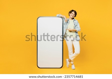 Full body smiling young woman she wear green t-shirt denim shirt casual clothes point index finger on big huge blank screen mobile cell phone smartphone with area isolated on plain yellow background