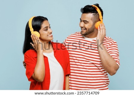 Young happy couple two friends family Indian man woman wear red casual clothes t-shirt together listen music in headphones look to each other isolated on pastel plain light blue cyan color background