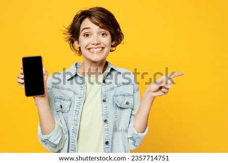 Young surprised woman wear green t-shirt denim shirt casual clothes hold in hand use mobile cell phone with blank screen workspace area point aside on area isolated on plain yellow background studio
