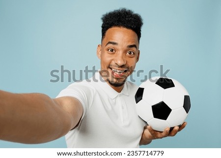 Close up young man fan wear t-shirt do selfie shot pov on mobile cell phone cheer up support football sport team hold soccer ball watch tv live stream isolated on plain pastel blue color background