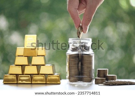 Woman hand putting a coin into glass bottle and gold bar on natural green background,Business investment and Saving money for prepare in future concept