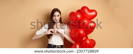 Image of beautiful young woman smiling and showing heart gesture, I love you sign, standing near romantic balloons on Valentines day, beige background.