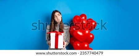 Valentines holiday concept. Pensive young woman in dress, holding gift and looking at upper left corner, thinking about lovers day, standing near red hearts balloons, blue background.