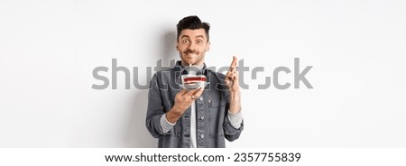 Birthday. Happy guy making wish with fingers crossed, holding bday cake with candle, standing on white background.