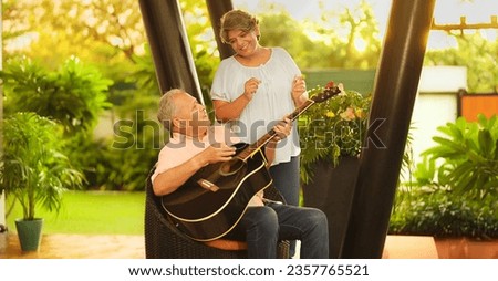 Happy mature middle aged man guitarist sitting on chair play acoustic guitar enjoy music outdoor home. Beautiful smiling Indian female singing practice tuning on musical instrument spend time together Royalty-Free Stock Photo #2357765521