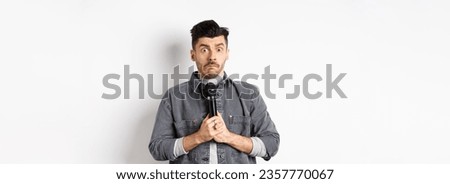 Nervous unconfident guy holding microphone and look indecisive, scared to perform, standing shy against white background.