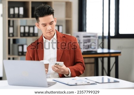 Happy young man looking at cellphone screen, reading message, or email, communicating distantly with client from tablet computer work in modern office.