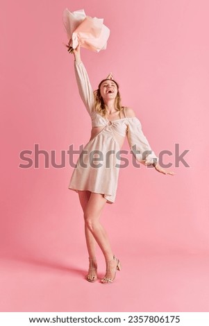 Full-length image of happy emotional young woman in cute dress standing with flowers and laughing over pink studio background. Concept of human emotions, fashion, youth, party, celebration, sales, ad