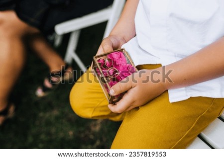 A child, a boy, holds in his hands a glass box with flowers, roses, golden rings with words, an inscription about love forever at a wedding ceremony. Photo close-up, portrait.