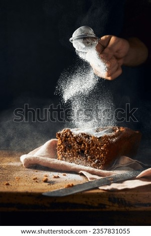 Sliced banana bread on a rustic wooden table and black background. Cropped hand is dusting sugar powder on top of it. Vertical still life picture. Space for copy.