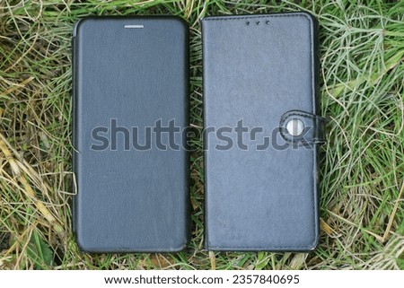 two smartphones in black leather closed cases lie in the green grass on the street