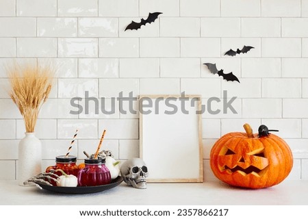 Carved Halloween pumpkin, picture frame mockup, vase of dry flowers, drinks, decorations on table in scandinavian kitchen. Happy Halloween concept.