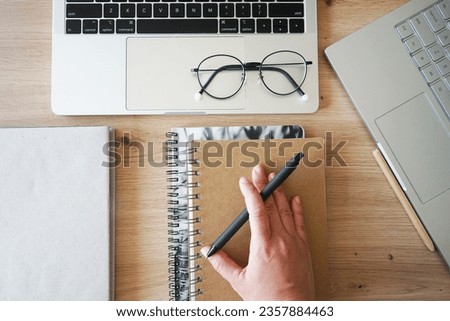 A hand holds a pen on a notepad next to a book and two open notebooks on a wooden table