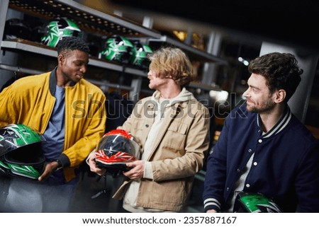 happy multicultural male friends in jackets holding helmets, indoor racing track, karting concept Royalty-Free Stock Photo #2357887167